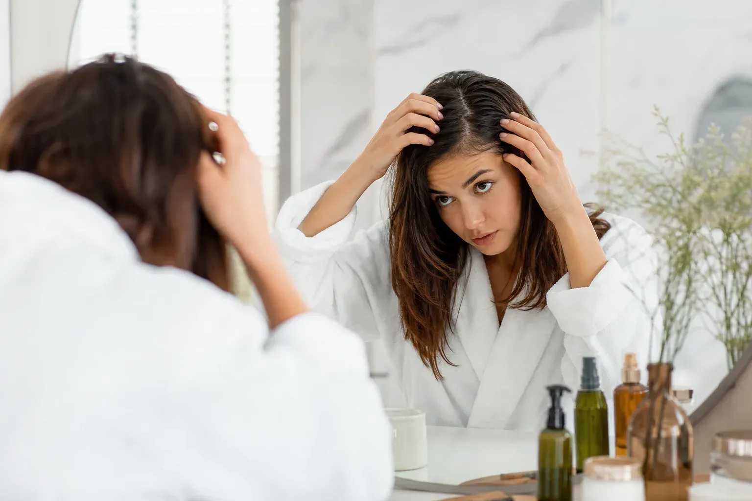 Femme en peignoir examinant ses racines de cheveux devant un miroir de salle de bain